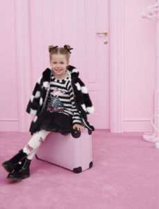 bambina sorridente seduta su valigia con abiti bianco e nero in una stanza completamente rosa
